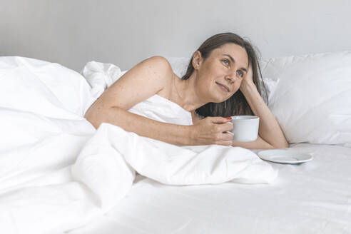 Nachdenkliche Frau, die eine Kaffeetasse hält und zu Hause im Bett liegt - EVKF00060