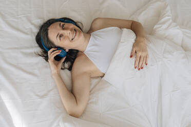 Smiling woman wearing wireless headphones listening to music in bed - EVKF00062