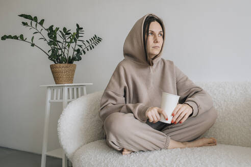 Contemplative woman sitting with disposable cup on sofa - EVKF00072