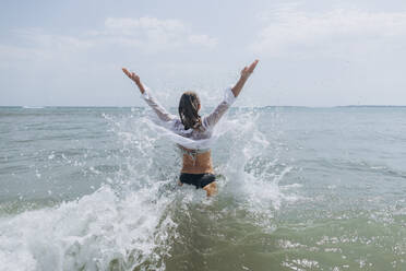 Frau mit erhobenen Armen stehend und Wasser spritzend im Meer - SIF00900