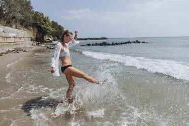 Playful woman splashing water with leg at beach - SIF00902