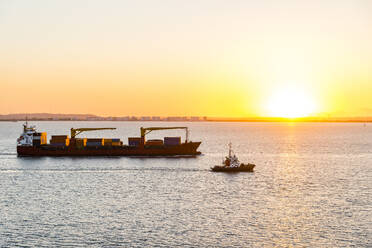 Spain, Andalusia, Cadiz, Boat sailing past moving container ship at sunset - EGBF00939