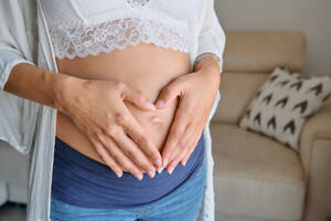 Nicht erkennbare schwangere Frau in weißem BH und Jeans, die zärtlich ihren Bauch berührt, während sie in der Nähe des Sofas im heimischen Wohnzimmer steht - ADSF46691