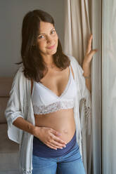Junge schwangere Frau in weißem BH und Jeans lächelnd und streichelnden Bauch mit Zärtlichkeit, während stehend in der Nähe von Fenster im Wohnzimmer zu Hause - ADSF46695