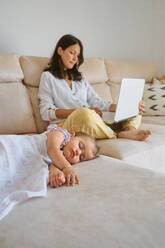Ganzkörperdarstellung einer jungen Frau, die auf einem gemütlichen Sofa in der Nähe ihrer schlafenden Tochter im heimischen Wohnzimmer auf ihrem Laptop surft - ADSF46696
