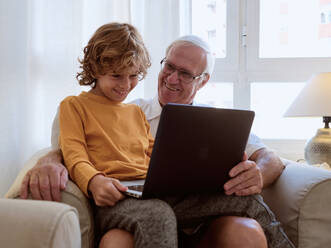 Cheerful senior man looking at positive grandson surfing modern netbook while sitting in comfortable armchair in living room at home - ADSF46704