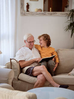 Lächelnder Enkel in Freizeitkleidung, der seinen Großvater anschaut, während er gemeinsam auf einer bequemen Couch im hellen Wohnzimmer zu Hause ein Buch liest - ADSF46705