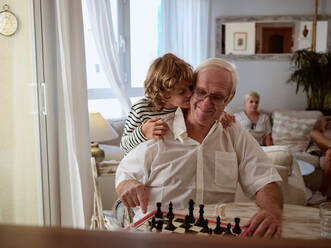 Positiver Junge, der seinen zufriedenen Großvater umarmt, der am Tisch mit einem Schachbrett sitzt, während er zusammen im hellen Wohnzimmer zu Hause spielt - ADSF46707