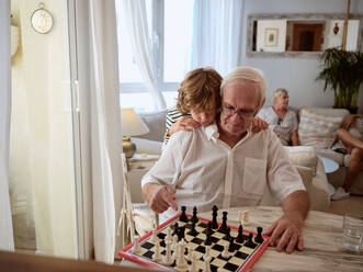 Preteen kid embracing senior man and learning to play chess at table in cozy living room at home - ADSF46709
