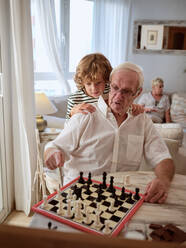 Focused preteen boy hugging elderly grandfather while learning to play chess at table in living room at home - ADSF46713