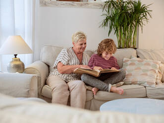 Positive elderly woman sitting near grandson and showing interesting moment and talking existed story in living room with cozy interior - ADSF46715