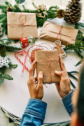 High angle of crop unrecognizable person holding gift box above white decorated table with Christmas presents - ADSF46721