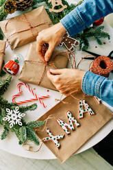 Von oben von Ernte unerkennbar Person Verpackung Weihnachtsgeschenke in Papier mit Seil am Tisch mit Tannenzweigen und Glas Schneekugel in der Nähe von Buchstaben und Zuckerstangen - ADSF46724