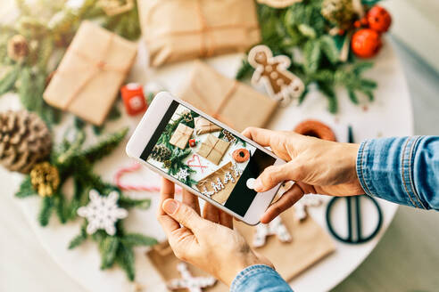 Overhead-Ansicht der Ernte anonyme Person, die ein Foto von eingepackten Weihnachtsgeschenken auf dem Handy in der Nähe von Tannenzweigen und Papier neben dem Wort xmax und Zuckerstangen - ADSF46726