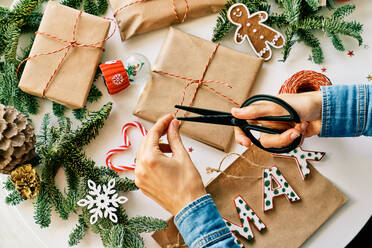 Draufsicht auf eine anonyme Person, die Weihnachtsgeschenke in Papier einwickelt, während sie ein Seil mit einer Schere an einem Tisch mit Fichtenzweigen und einer gläsernen Schneekugel in der Nähe von Buchstaben und Dekorationen schneidet - ADSF46727