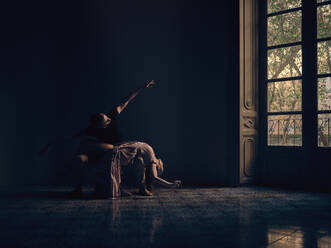Full body of ballerina lying on knees of male dancer while performing ballet dance movement in dark room with window - ADSF46729