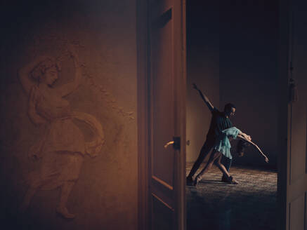 Seitenansicht eines anonymen Mannes und einer zarten Ballerina, die in einem leeren Raum hinter einer geöffneten Tür bei gedämpftem Licht tanzen - ADSF46730
