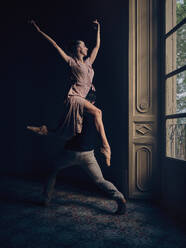 Full body side view of anonymous talented male dancer carrying graceful ballerina on shoulder while dancing ballet dance together in obscure room - ADSF46732