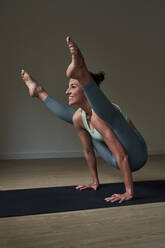 Ganzer Körper einer lächelnden Sportlerin in Sportkleidung, die die Tittibhasana-Pose auf einer schwarzen Matte einnimmt, während sie in einem modernen Club Yoga übt - ADSF46737