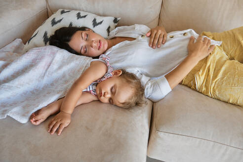 From above of young mother and cute little baby covered with blanket sleeping together on sofa in living room at home - ADSF46747