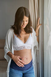 Friedliche Frau in weißer Unterwäsche und Jeans schließt die Augen, während sie zu Hause am Fenster steht und den schwangeren Bauch berührt - ADSF46748