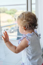 Side view of curious baby girl leaning on window and exploring street outside of house while standing in living room at home - ADSF46753