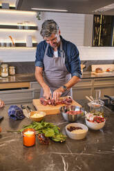 Cheerful elder male in apron cutting meat for tasty dish on wooden board in modern kitchen - ADSF46755