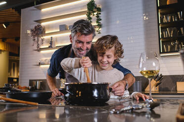 Glücklicher Junge mit lockigem Haar, der ein heißes Gericht in einem Kochtopf umrührt und seinem Vater im mittleren Alter beim Kochen hilft, während er seine Zeit in der Küche zu Hause verbringt - ADSF46759