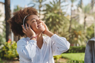 Lächelnde junge afroamerikanische Frau mit Kopfhörern, die mit geschlossenen Augen im Stadtpark an einem hellen Sommertag Musik hört - ADSF46760