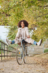 Full length of playful African American woman riding bicycle with raised legs while having fun in autumn park - ADSF46761