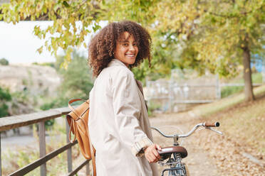 Side view of young African American female with curly hair in stylish coat standing with bicycle and looking at camera in park - ADSF46762