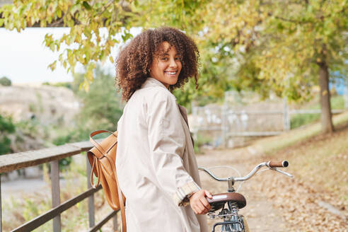 Seitenansicht einer jungen Afroamerikanerin mit lockigem Haar in einem eleganten Mantel, die mit einem Fahrrad im Park steht und in die Kamera schaut - ADSF46762