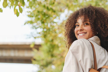 Von unten Seitenansicht der positiven afroamerikanischen Frau mit lockigem Haar und zahnigem Lächeln, die im Park wegschaut - ADSF46763
