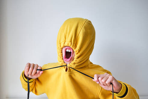 Anonymous child in yellow hoodie shouting and stretching drawstrings while tightening hood covering face against gray background - ADSF46768