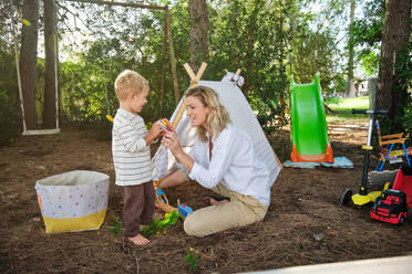 Full body of little boy and mother in casuals sitting on ground and playing with toy while spending time in nature - ADSF46770