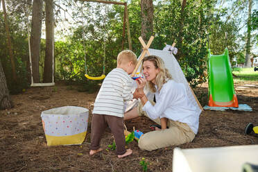 Full body of cheerful woman playing with little boy while sitting near tent in park - ADSF46774