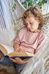 Konzentrierter Junge mit lockigem Haar, der eine interessante Geschichte liest, während er sich in einer gestrickten weißen Hängematte auf dem Balkon zu Hause ausruht - ADSF46777