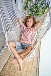 From above full length of adorable barefoot boy in casual outfit relaxing in wicker hammock with closed eyes and crossed legs on balcony with rug - ADSF46779