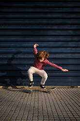 Full body energetic boy in casual clothes doing trick on skateboard on pavement against dark blue wall in weekend on city street - ADSF46783