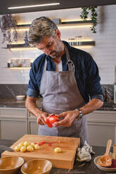 Concentrated bearded man in apron standing near kitchen counter and peeling tomato over chopping board - ADSF46808