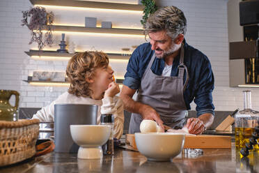 Optimistic man and son looking at each other while cutting fresh onion at table with various dishware in light kitchen - ADSF46809