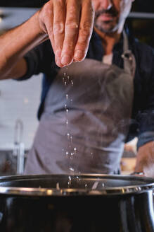 Mann in grauer Schürze hält die Hand über einem Kochtopf und gibt eine Handvoll Salz ins Wasser - ADSF46810