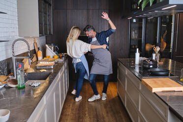 Back view of young woman hugging unshaven Hispanic man and dancing together in aprons while cooking food at home - ADSF46811