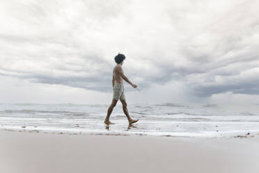 Mann läuft barfuß am Strand unter bewölktem Himmel - PBTF00247