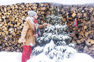 Senior woman decorating Christmas tree in front of firewoods - HHF05929