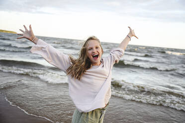 Cheerful girl standing with arms outstretched in front of sea - EYAF02818