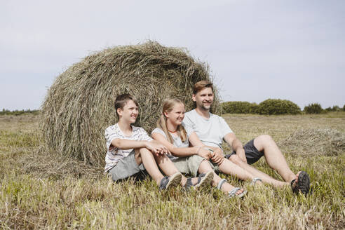 Lächelnde Kinder sitzen mit ihrem Vater in der Nähe von Heu auf einem Feld - EYAF02829