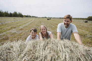 Happy father and children pushing and playing with hay in field - EYAF02831