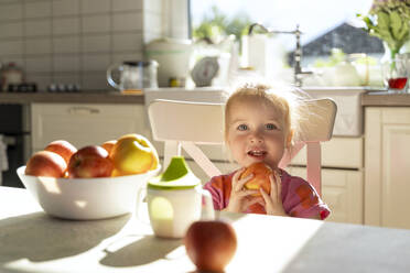 Glückliches Mädchen hält Apfel Obst sitzen am Esstisch in der Küche - NDEF01150