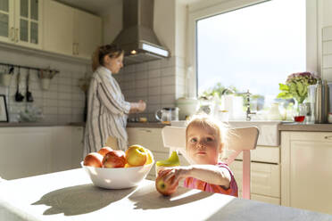 Mädchen hält Apfel am Esstisch, während die Mutter in der Küche kocht - NDEF01151